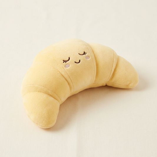 A soft plush crescent moon pillow in pale yellow with embroidered closed eyes, a smiling mouth, and small pink cheeks, photographed on a plain light beige background.
